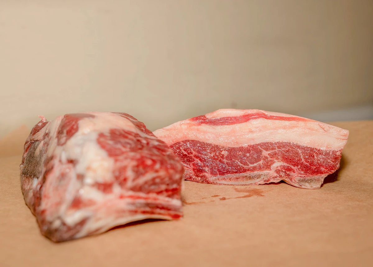 Two raw beef cuts with visible fat marbling, placed on a tan surface against a plain beige background.