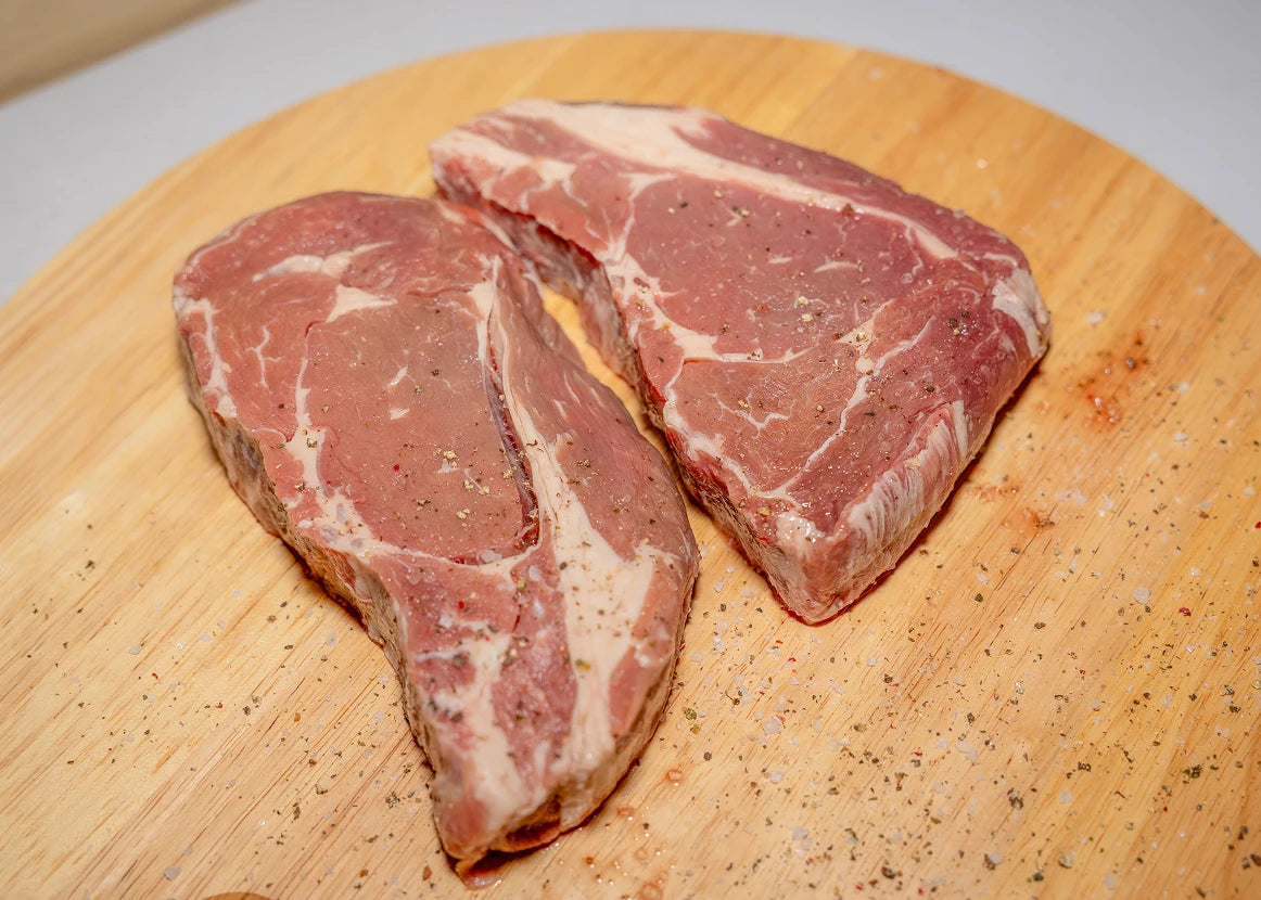 Two raw marbled beef steaks with visible seasoning, resting on a round wooden cutting board.